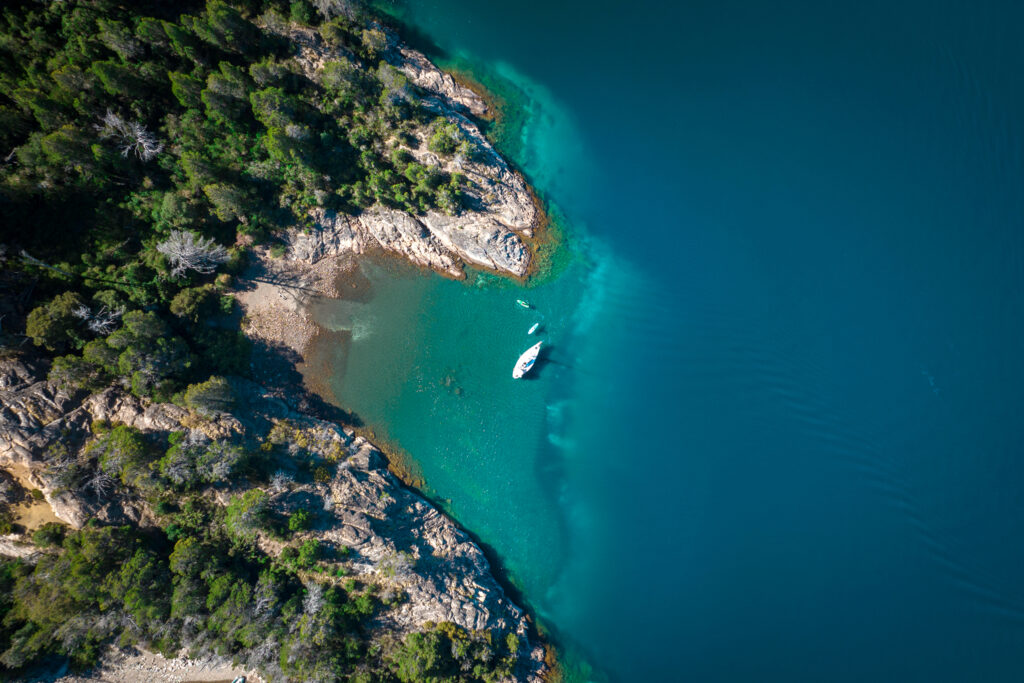 Vista aérea de um veleiro navegando nas águas turquesas do lago Nahuel Huapi, cercado por mata nativa, durante a experiência Sailing Tour Spirit x Rutini Wines