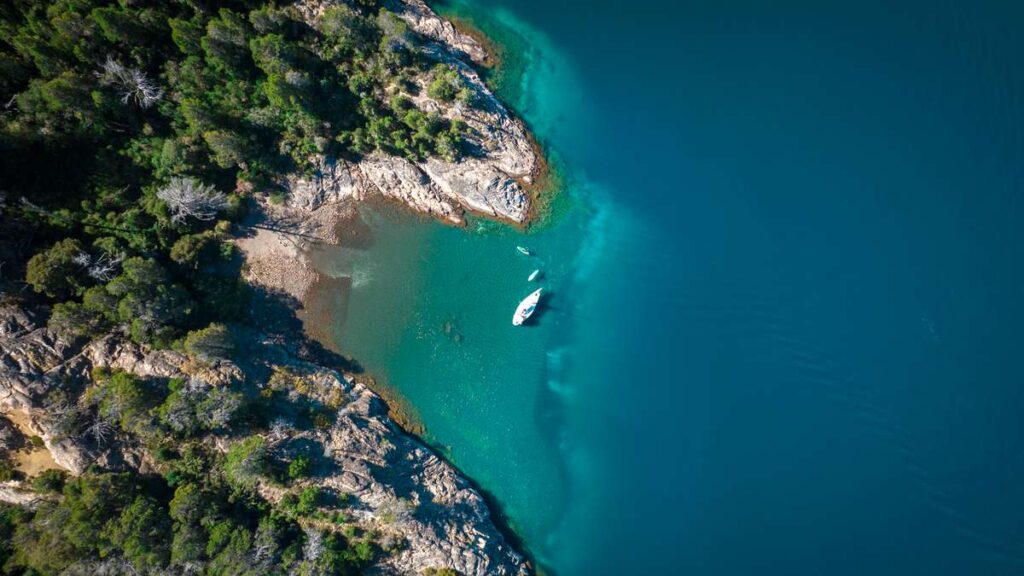 Vista aérea de un velero navegando en las aguas turquesas del lago Nahuel Huapi, rodeado de bosque nativo, durante la experiencia Sailing Tour Spirit x Rutini Wines