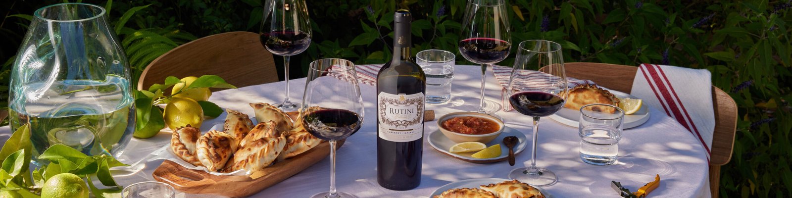 Bodega Rutini Wines - Colección y Maridaje Mesa con copa de vino tinto, botellas de vino Rutini Colección y platos con maridajes sobre un mantel de tela.